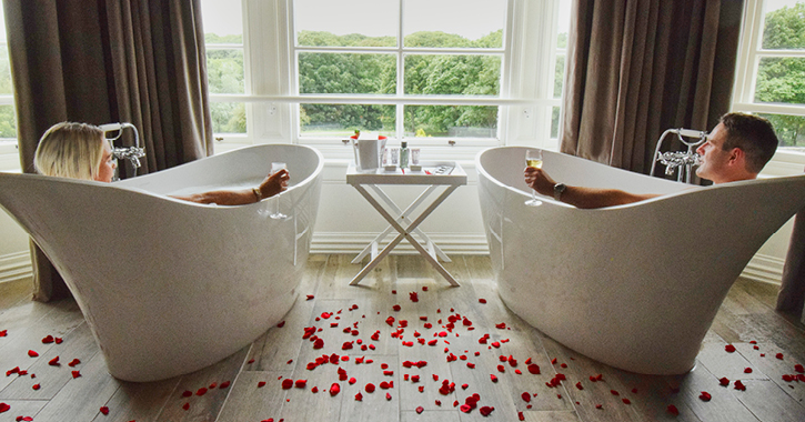 couple in matching bath tubs with rose petals on the ground in a hotel room at Seahm Hall. 